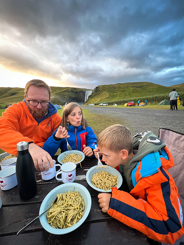 bambini con papà mangiano le linguine al pesto in un campeggio con una cascata sullo sfondo